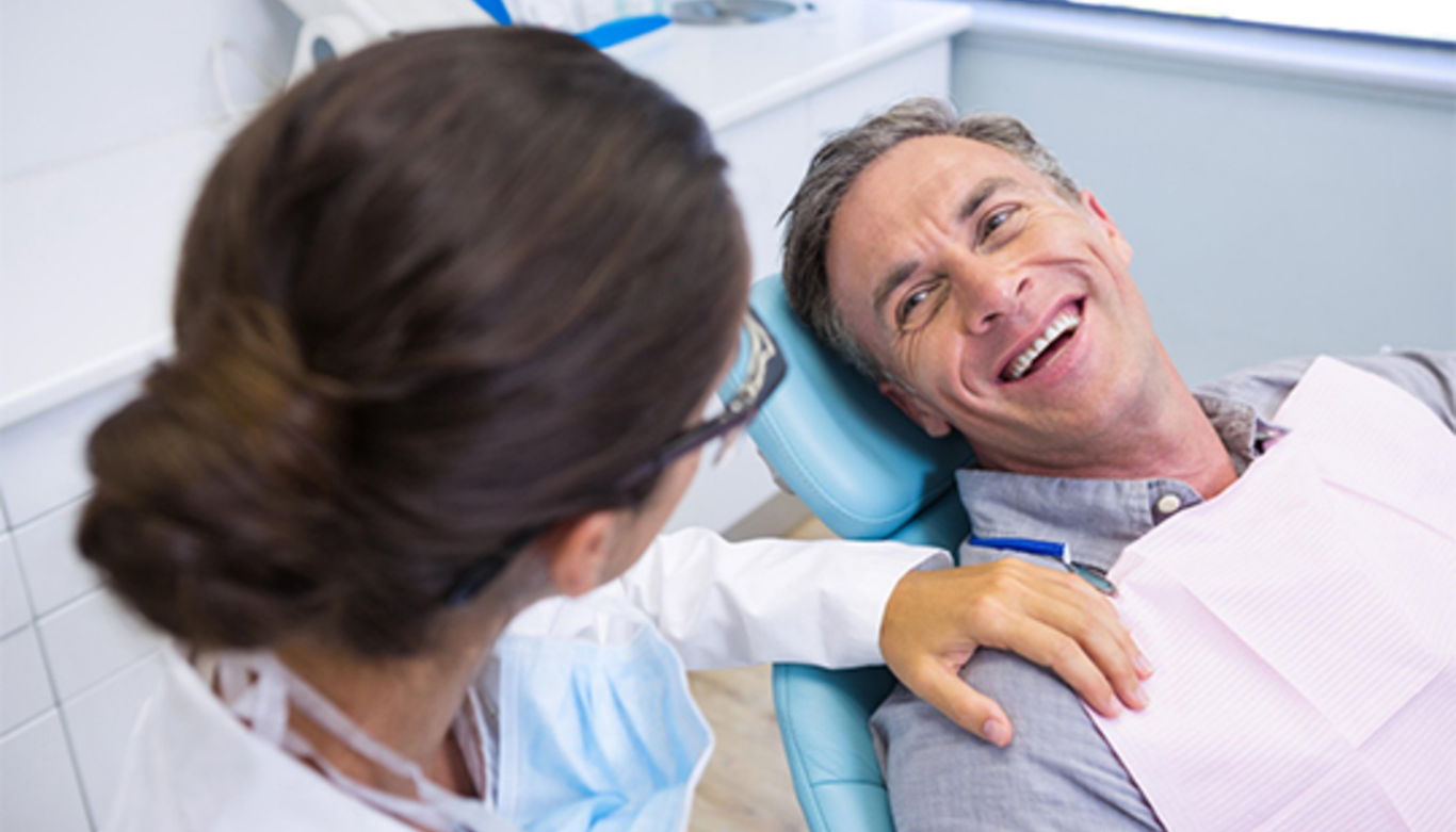 Dentist in white coat chatting with a smiling man in a dental chair during a checkup/smile visit.